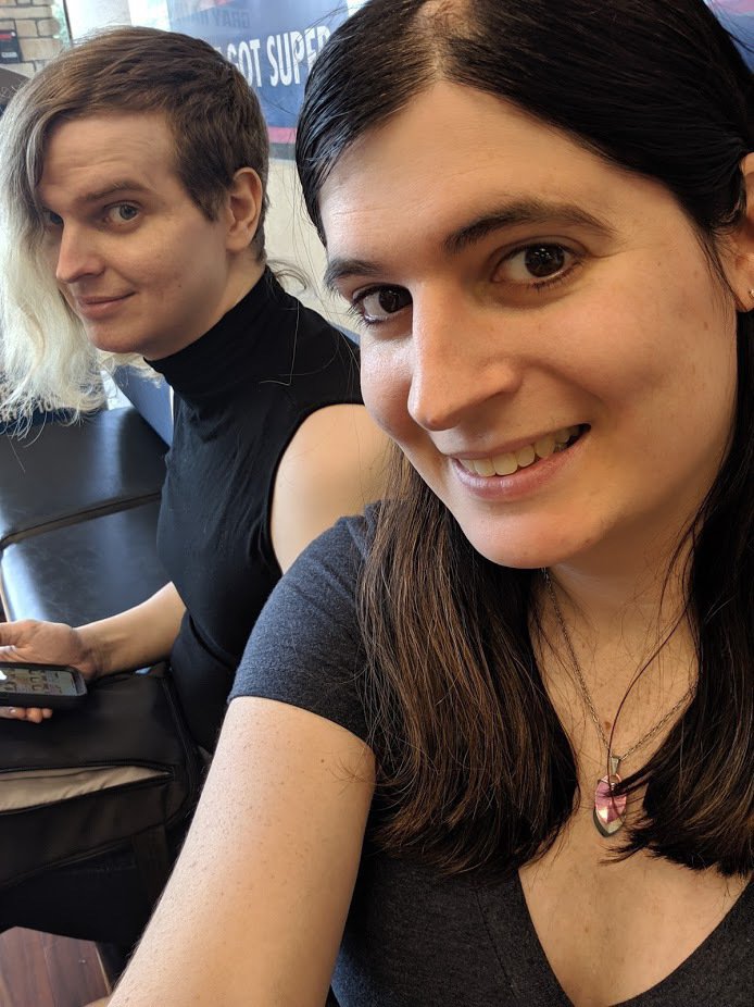 Selfie of Erika smiling in the foreground while Zoey, in a black sleeveless turtleneck, looks over from a bench behind her