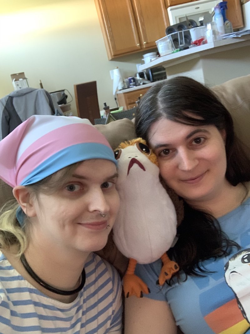 Zoey, wearing a trans pride flag bandana, and Erika press their cheeks against a plush porg between them