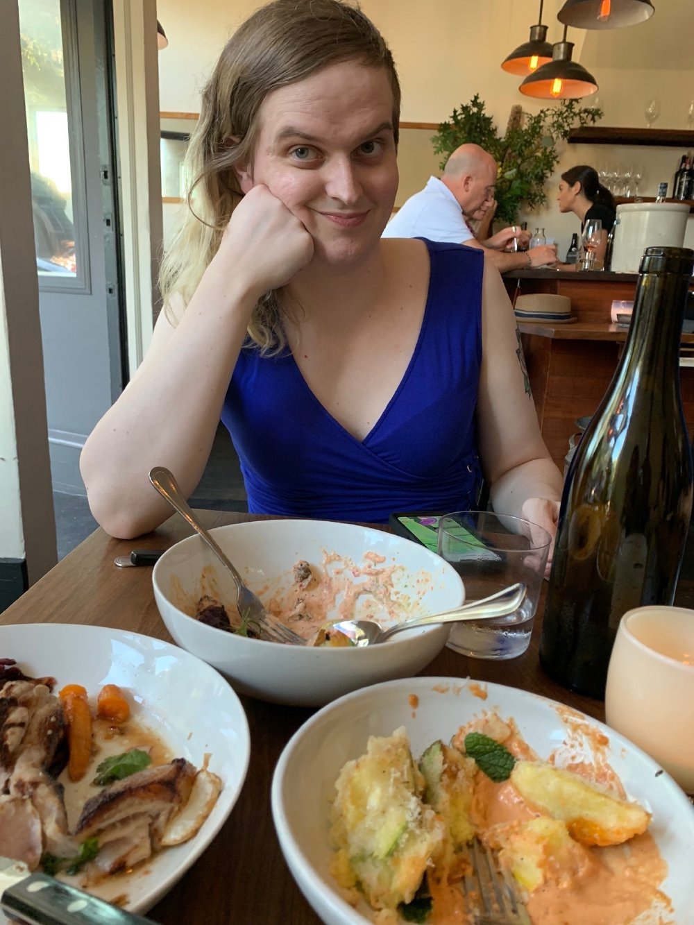 Zoey, in a blue dress at a restaurant table, smiling with her chin resting on her hand